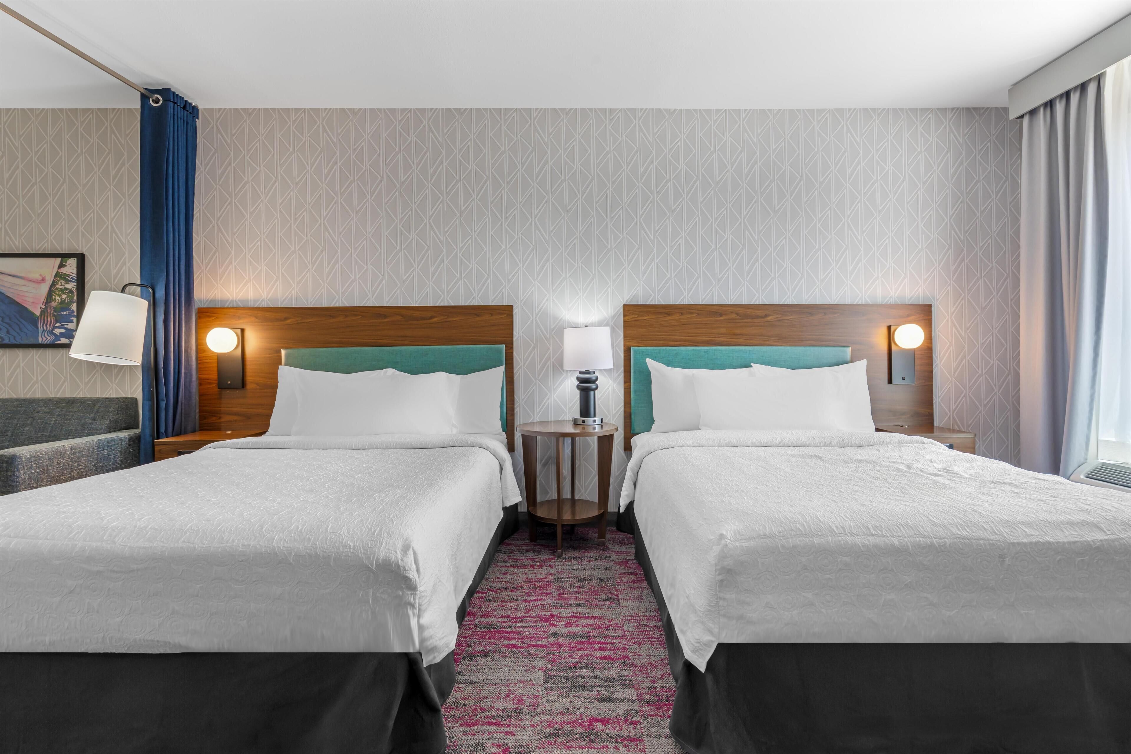A hotel room with two beds, a small table, a lamp, and a painting on the wall.
