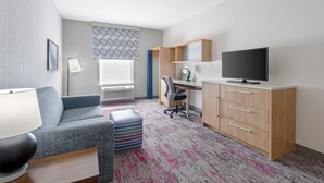 A modern hotel room with a sofa, a desk, a television, and a patterned rug. - Home2 Suites by Hilton Dallas DeSoto (DeSoto)