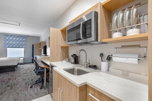 A modern hotel kitchen with stainless steel appliances, a sink, and a dining area with a chair and table. - Home2 Suites by Hilton Dallas DeSoto (DeSoto)