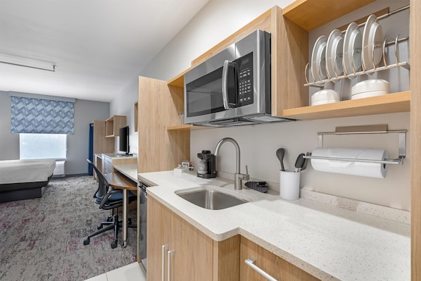 A modern hotel kitchen with stainless steel appliances, a sink, and a dining area with a chair and table. - Home2 Suites by Hilton Dallas DeSoto (DeSoto)
