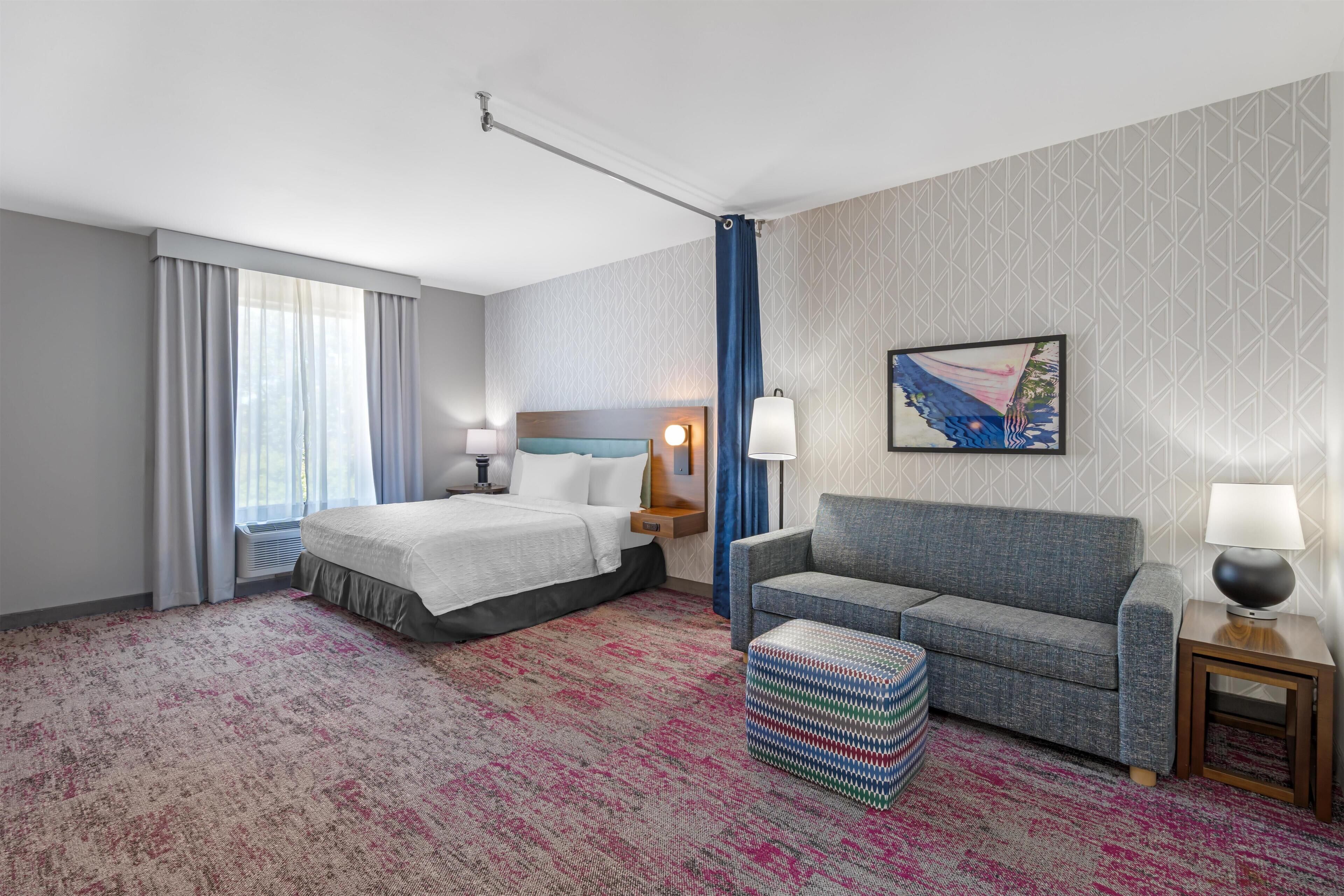 A hotel room with a bed, a sofa, a patterned carpet, a painting, and a window with curtains.