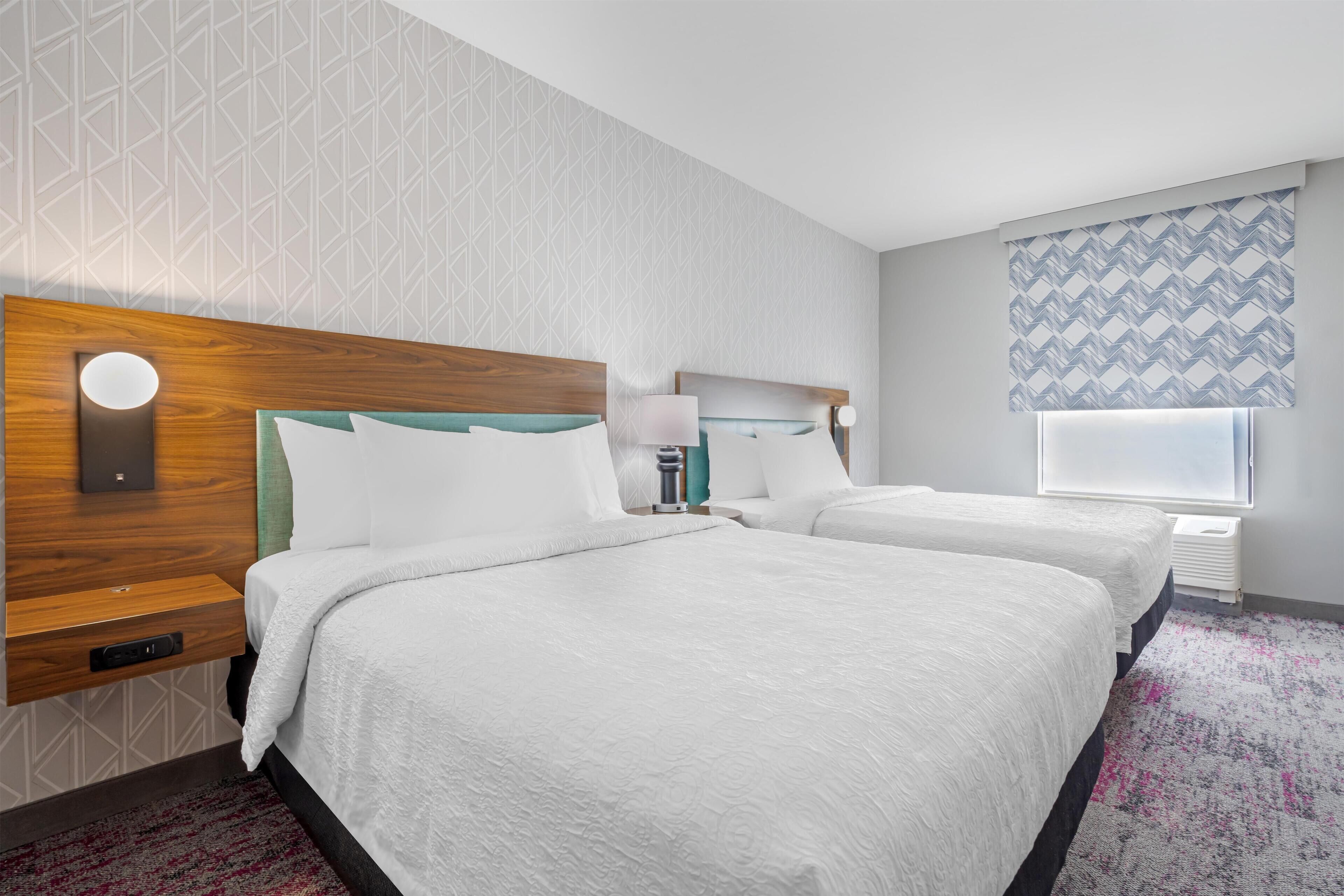 A hotel room with two beds, a wooden headboard, a nightstand, a lamp, and a window with patterned curtains.
