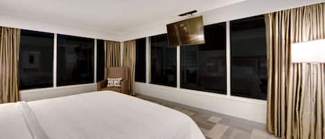 A hotel room with a large bed, a flat-screen TV mounted on the wall, and a view of the city through large windows.