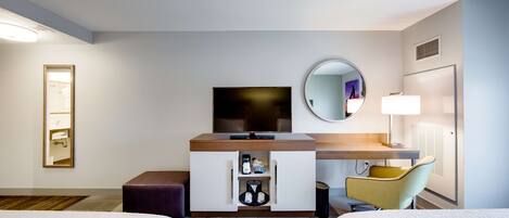 A hotel room with two beds, a TV, a minibar, a mirror, a desk with a lamp, and a chair.