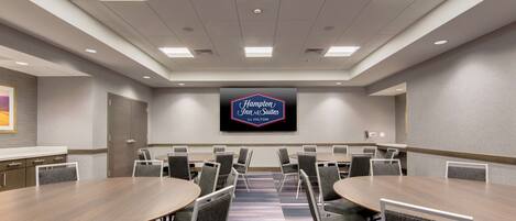 Un salón de conferencias con varias mesas y sillas, una pantalla grande mostrando el logotipo de Hampton Inn & Suites by Hilton, y piso alfombrado.