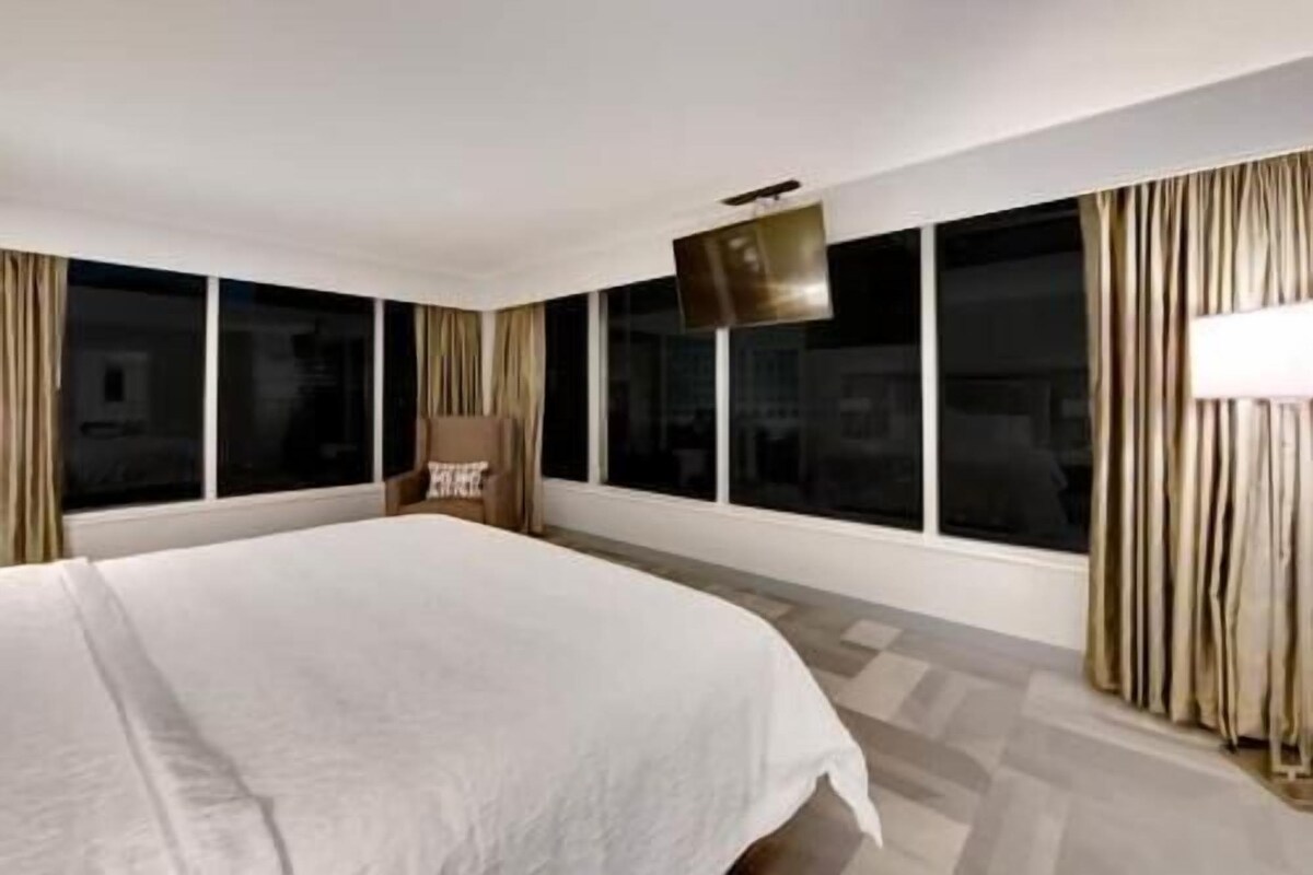A hotel room with a large bed, a flat-screen TV mounted on the wall, and a window with curtains.