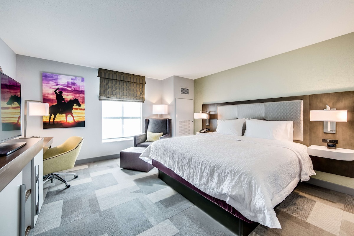 A modern hotel room with a large bed, a desk, a chair, and artwork on the walls.