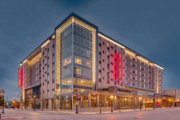 Un hotel moderno con fachada de vidrio, varios pisos y un llamativo letrero rojo.