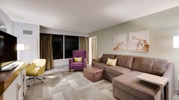 A modern hotel room with a sofa, armchair, desk, and a large window with curtains.
