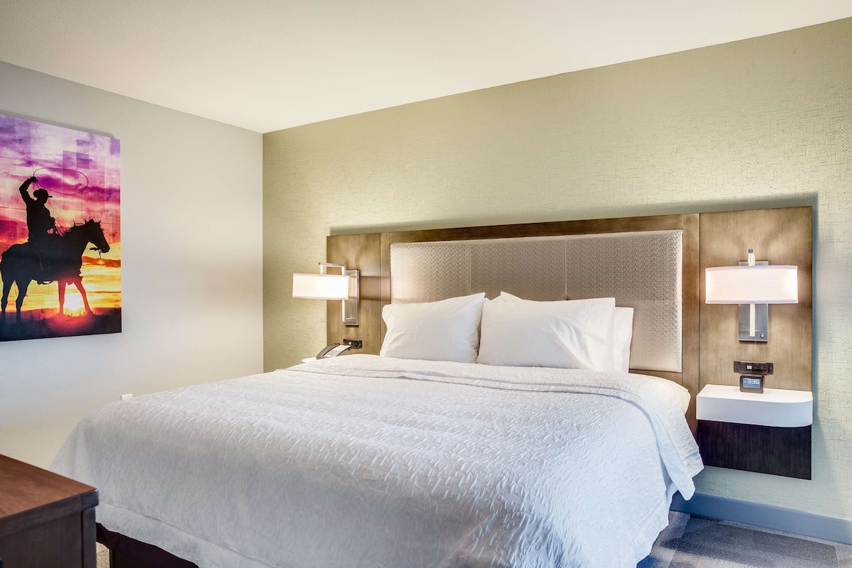 A hotel room with a large bed, two bedside lamps, a nightstand with a phone, and a painting of a cowboy on a horse.