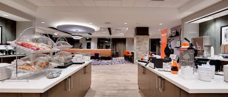 A hotel breakfast area with self-service stations, seating, and a view of the lobby.