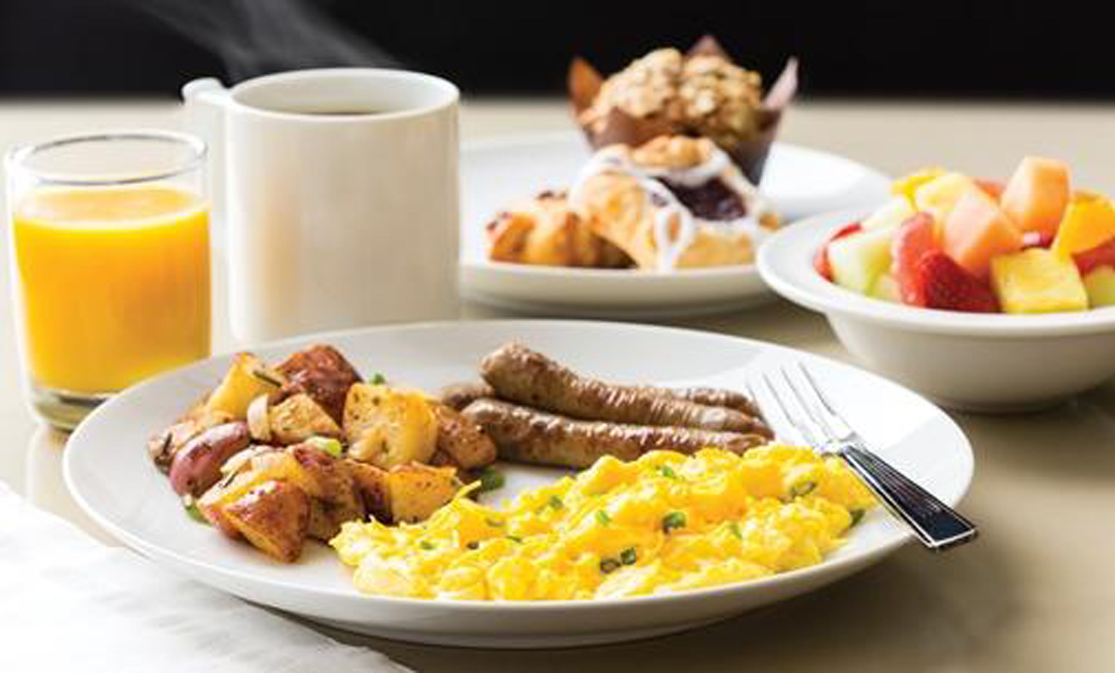 A breakfast plate with scrambled eggs, sausages, and potatoes, a glass of orange juice, a cup of coffee, a croissant, and a fruit bowl.