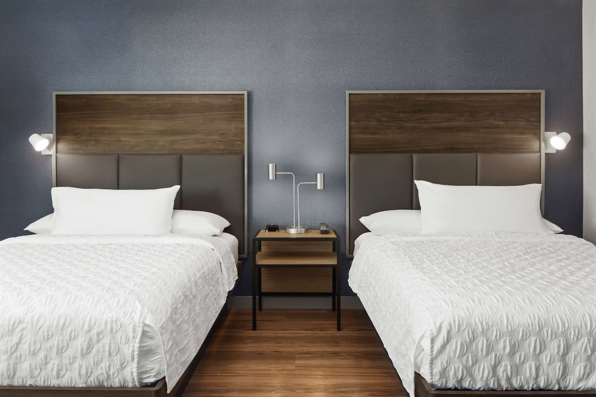 Une chambre d’hôtel avec deux lits, chacun doté d’une tête de lit en bois et de linge de lit blanc. Une petite table de chevet avec une lampe se trouve entre les deux lits.