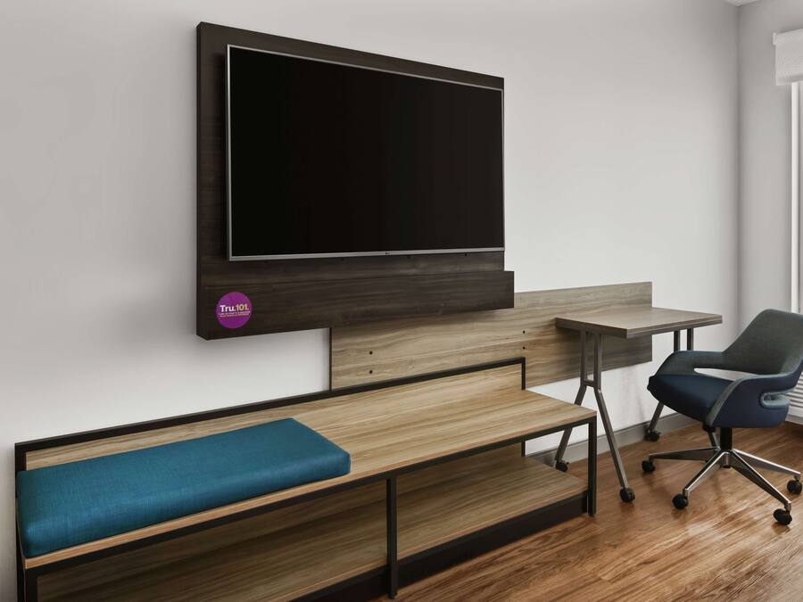 Una habitación moderna con una televisión de pantalla plana grande montada en la pared, un banco de madera con cojín azul, un escritorio con silla y una ventana que proporciona luz natural.