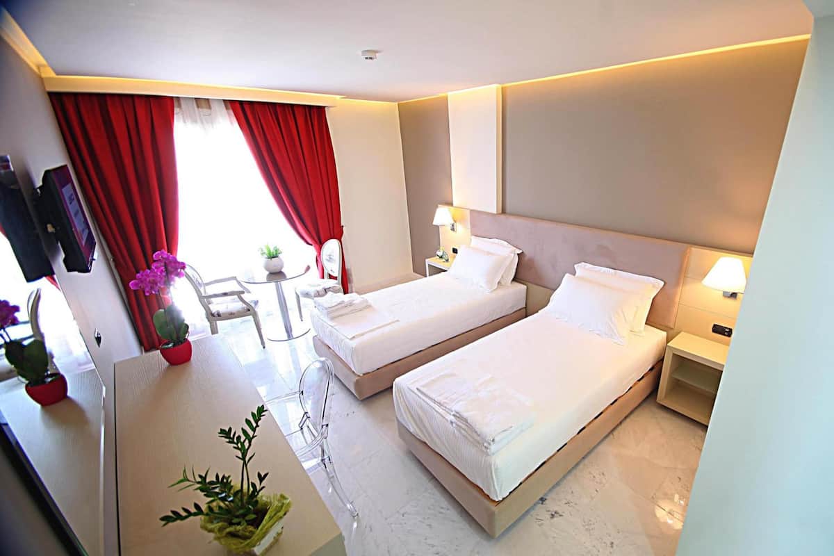 Quarto de hotel com duas camas, uma escrivaninha, uma cadeira, uma pequena mesa com uma planta e uma janela com cortinas vermelhas.