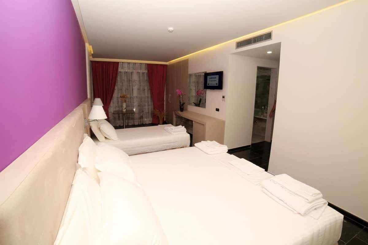 Quarto de hotel com duas camas, uma escrivaninha, um espelho e um banheiro visível por uma porta aberta.