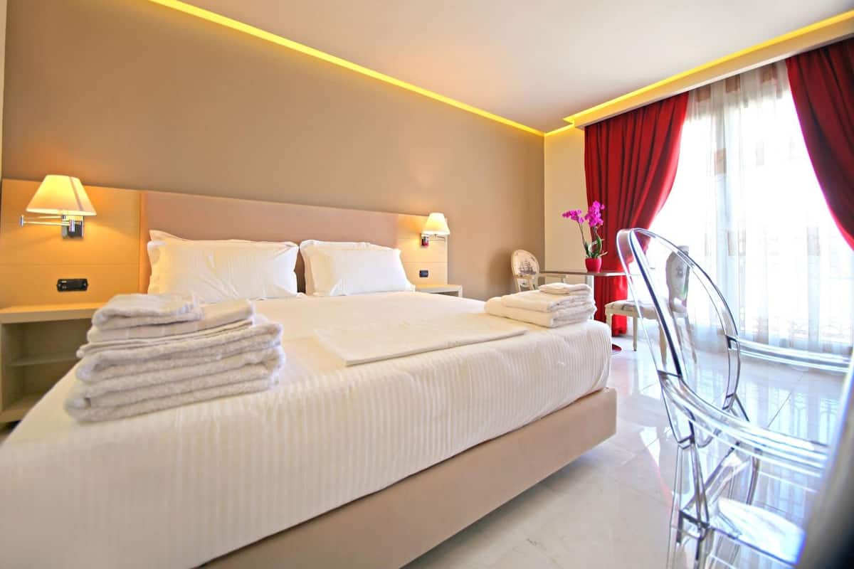 Quarto de hotel com uma cama grande, duas luminárias de cabeceira, uma cadeira e uma janela com cortinas vermelhas.