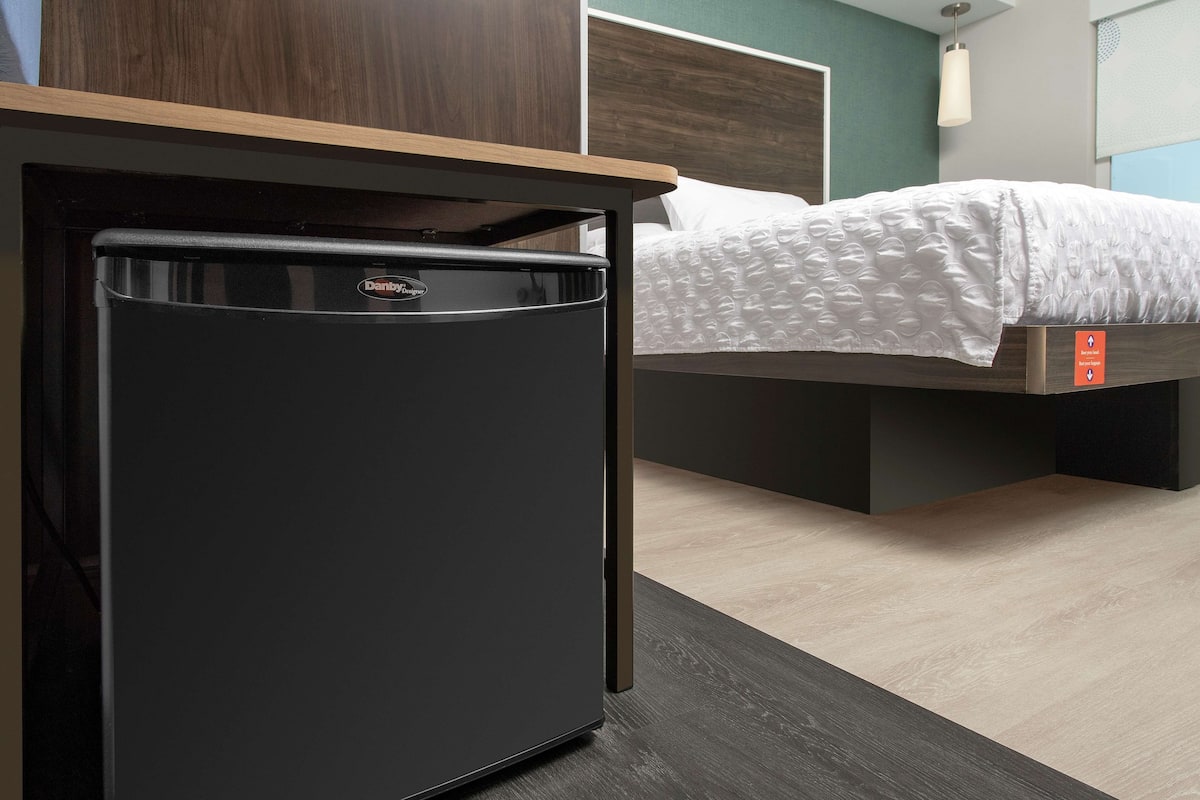 Ein kompakter, schwarzer Kühlschrank in einem Hotelzimmer mit Bett und Holz-Kopfteil.