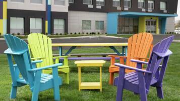 A set of colorful Adirondack chairs and a small yellow table arranged on a grassy area in front of a modern building with large windows and a blue awning.
