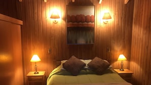 A room with wooden panel walls, a bed with green bedding and two pillows, two bedside lamps, and a painting above the bed. - Cabañas en Pucon con vista cordillera (Pucón)