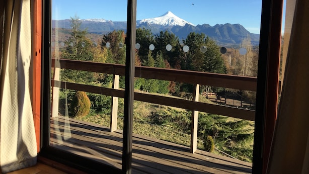 A view from a balcony with a wooden railing, overlooking a snow-capped mountain and a forested area. - Cabañas en Pucon con vista cordillera (Pucón)