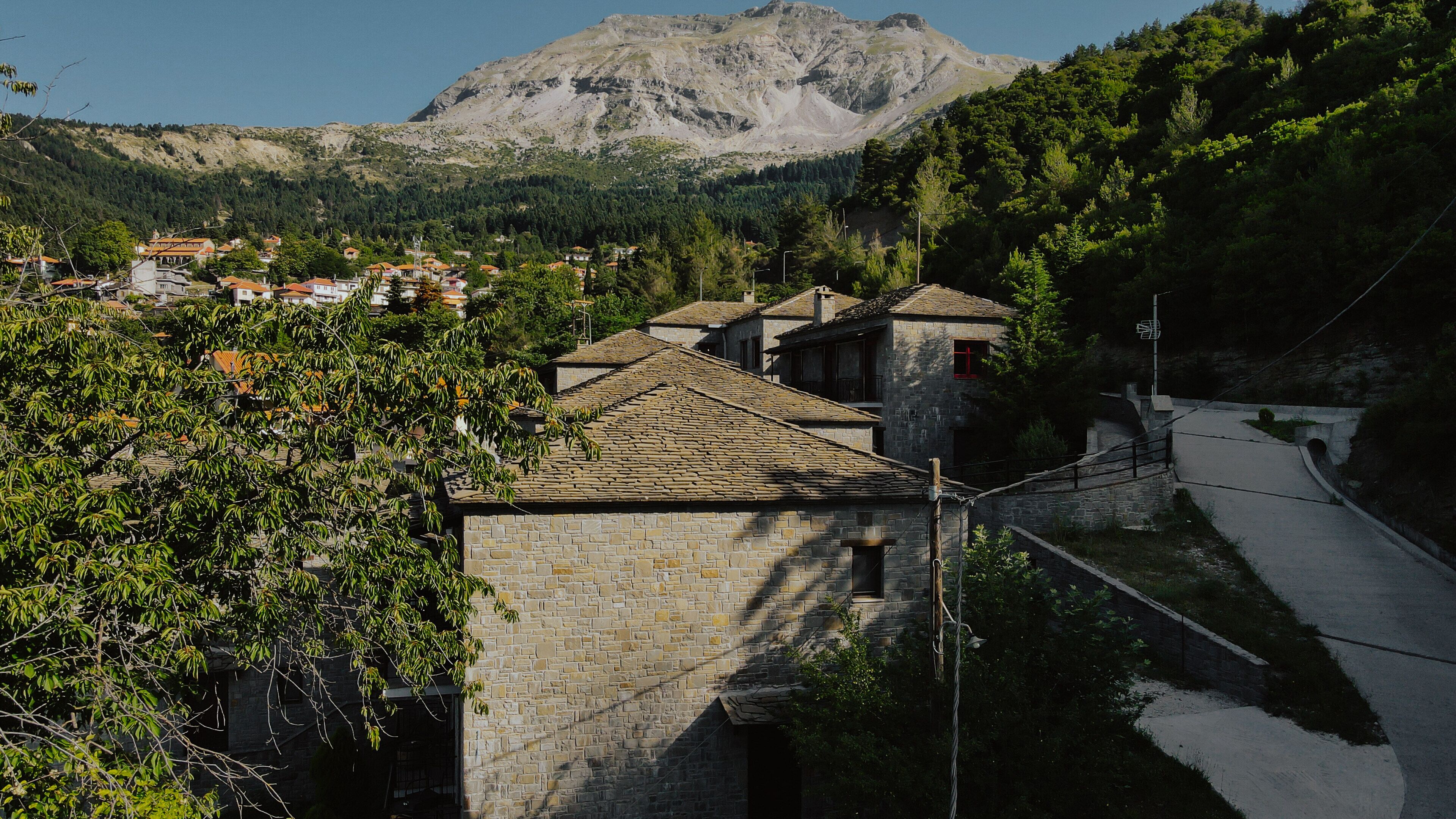 A stone building with a tiled roof, surrounded by trees and a mountain in the background.
