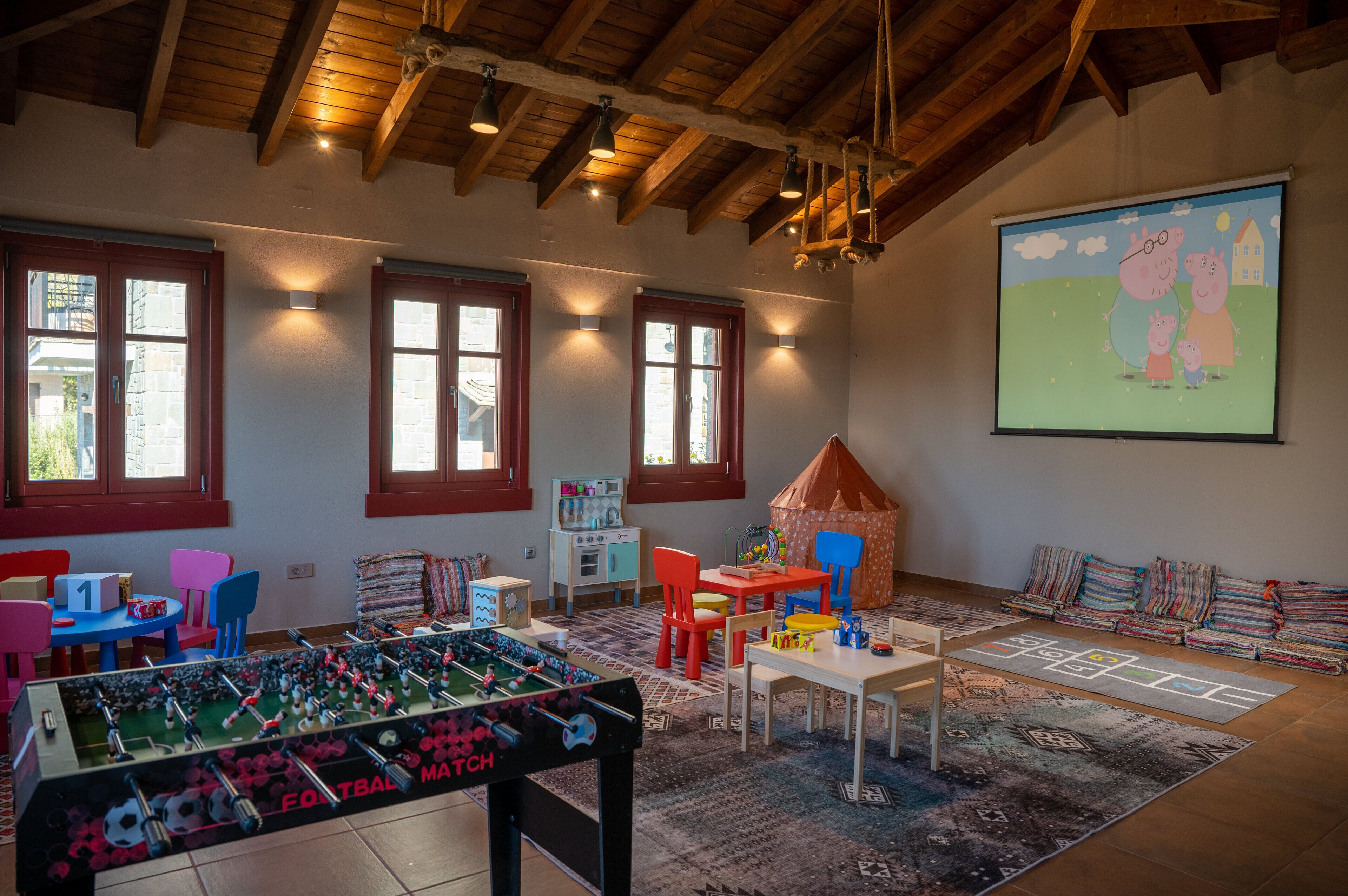 Een kamer met een tafelvoetbaltafel, kleurrijke stoelen, een kleintje tafeltje met speelgoed en een scherm waarop een tekenfilm wordt vertoond.
