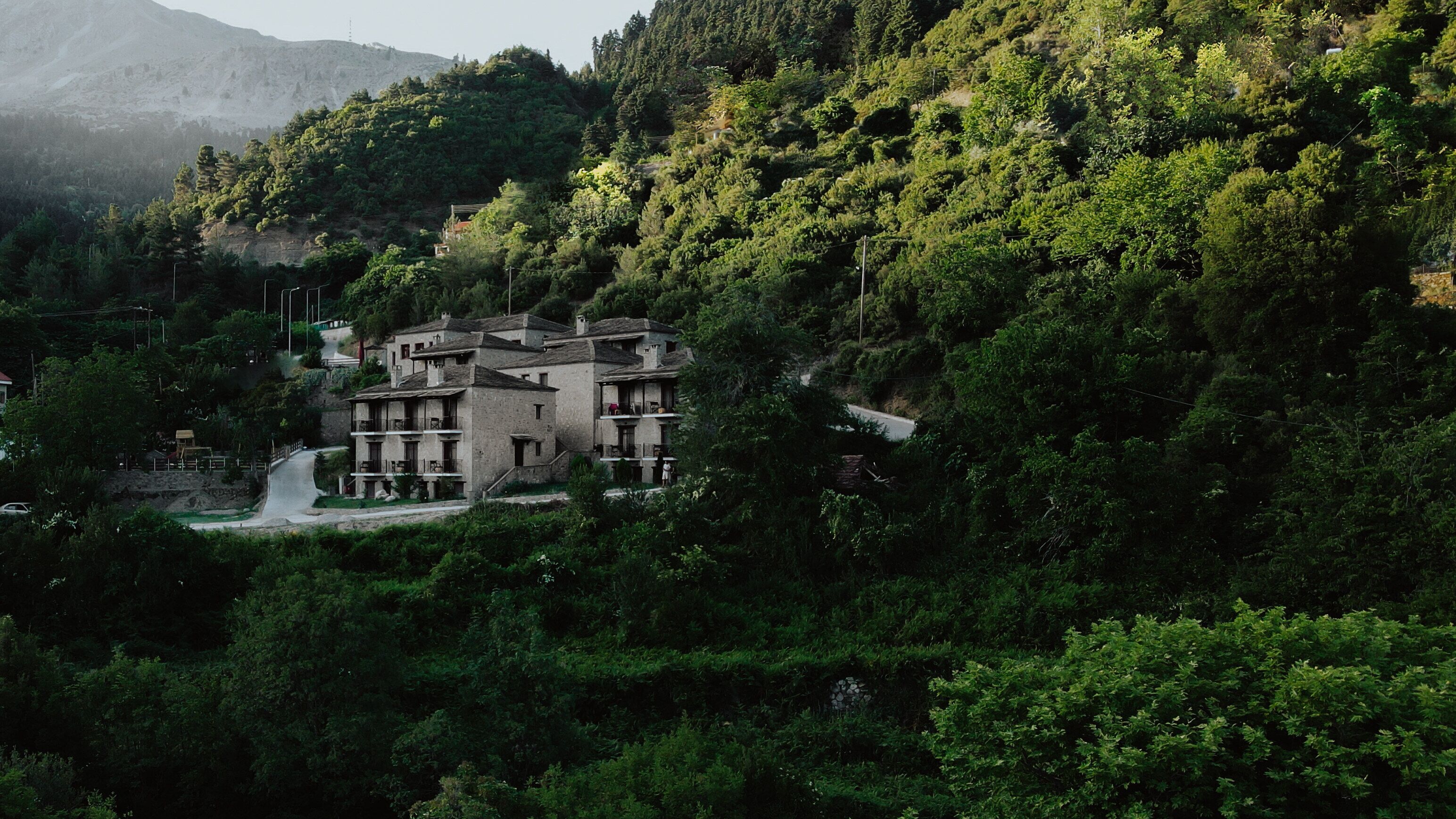 A traditional stone building with multiple balconies, surrounded by dense forest and a mountain in the background.
