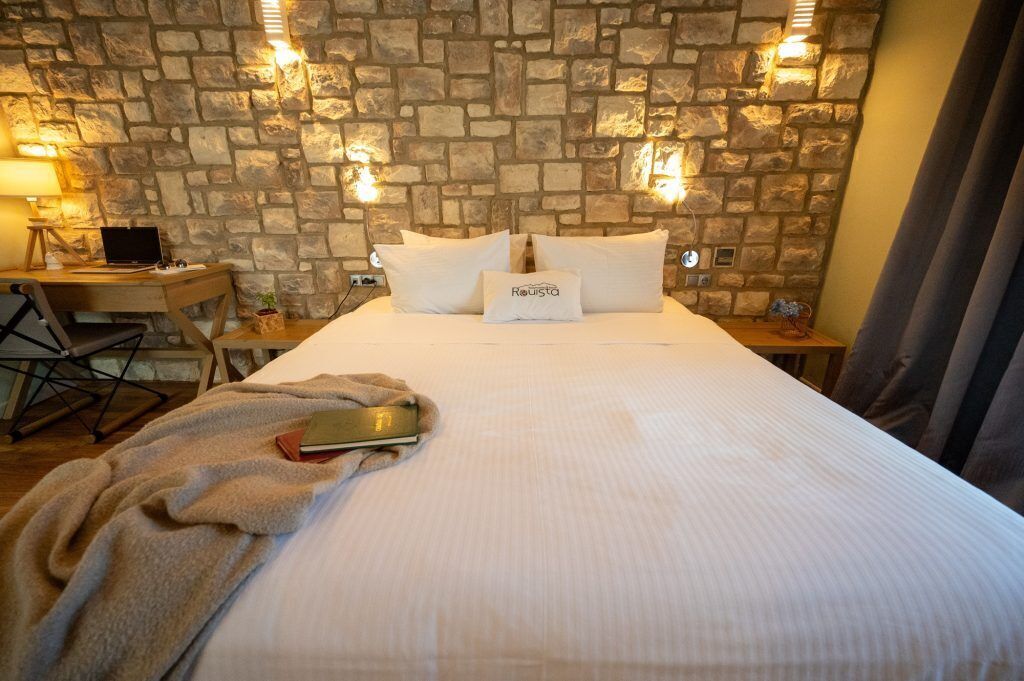 Habitación de hotel con pared de piedra, cama con ropa de cama blanca, escritorio con una computadora portátil y una manta en el suelo.