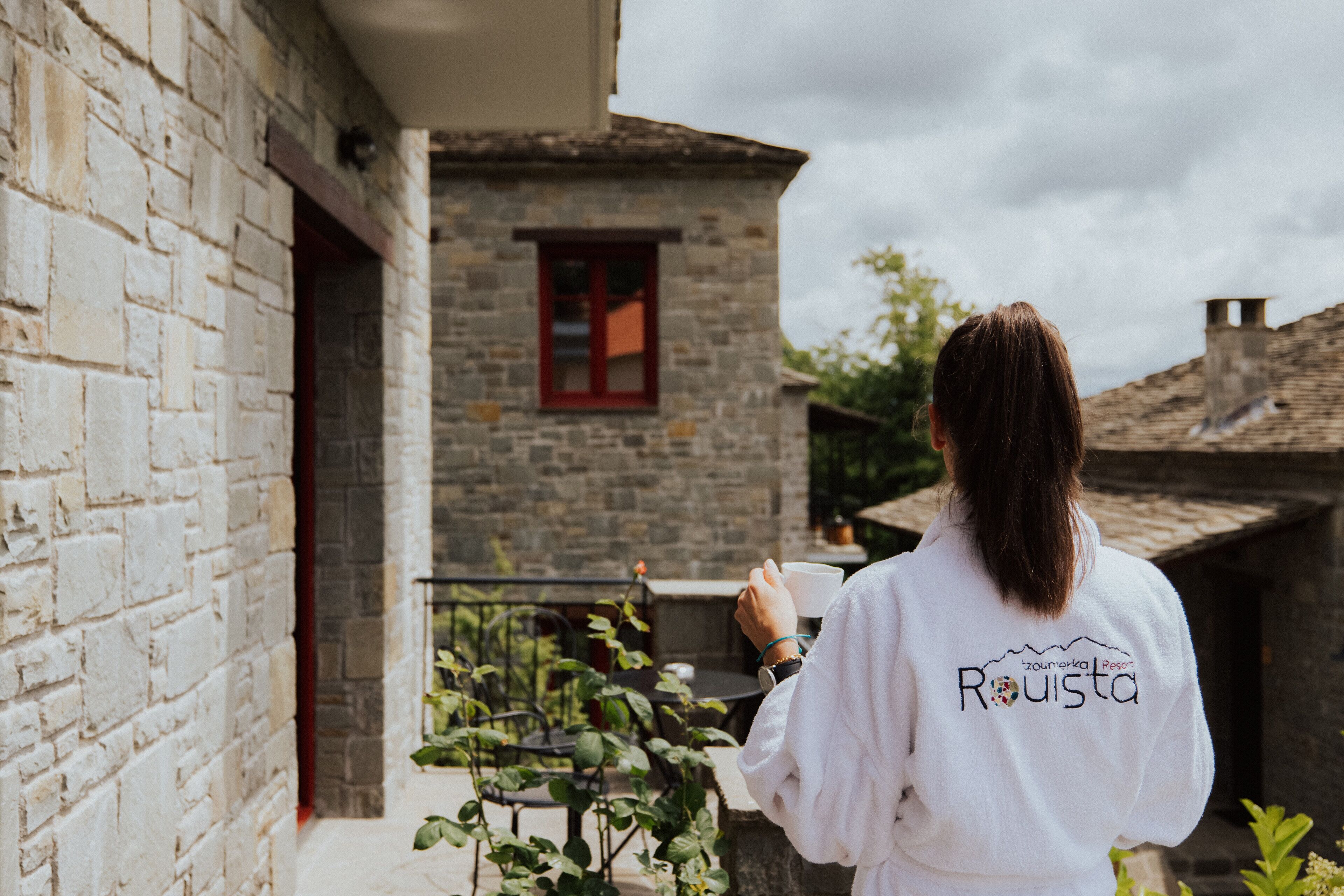 Una persona con una bata blanca que lleva el texto "Rousta", de pie en un patio con edificios de piedra y un ventanal rojo.