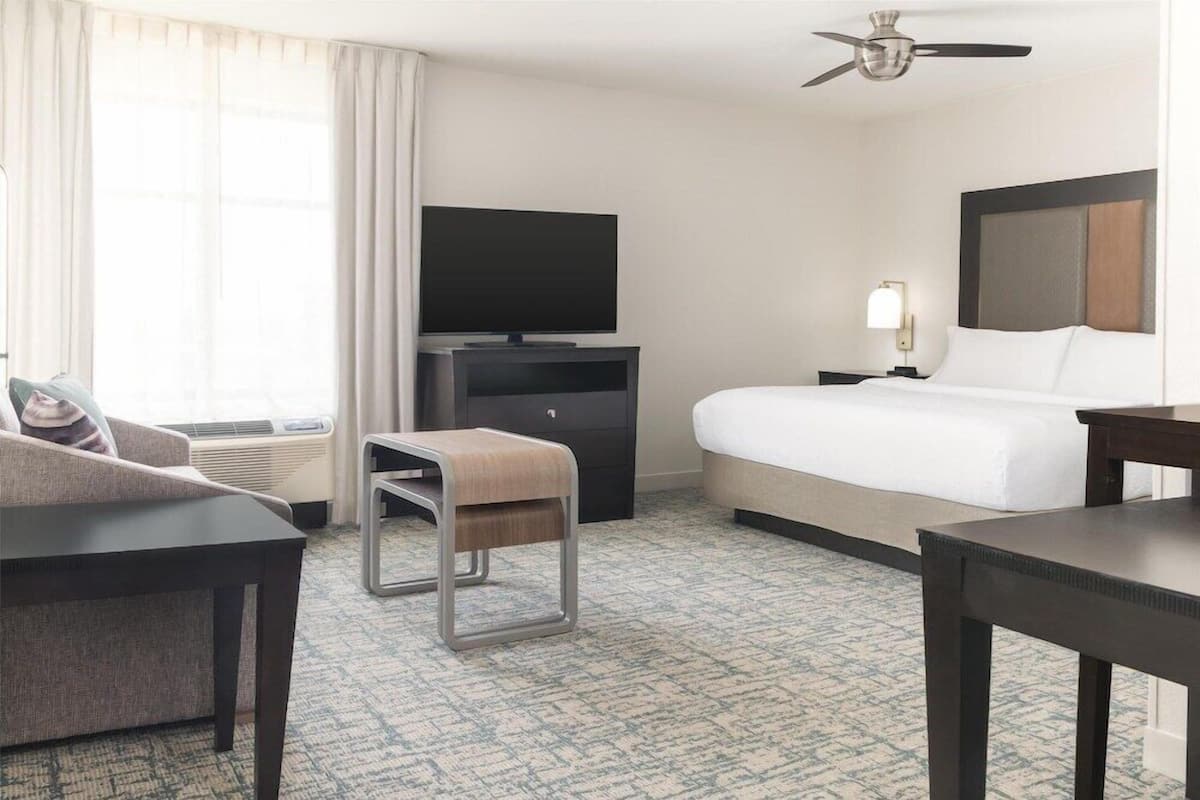 A hotel room with a bed, a television, a desk, a chair, a small stool, a nightstand with a lamp, and a ceiling fan.