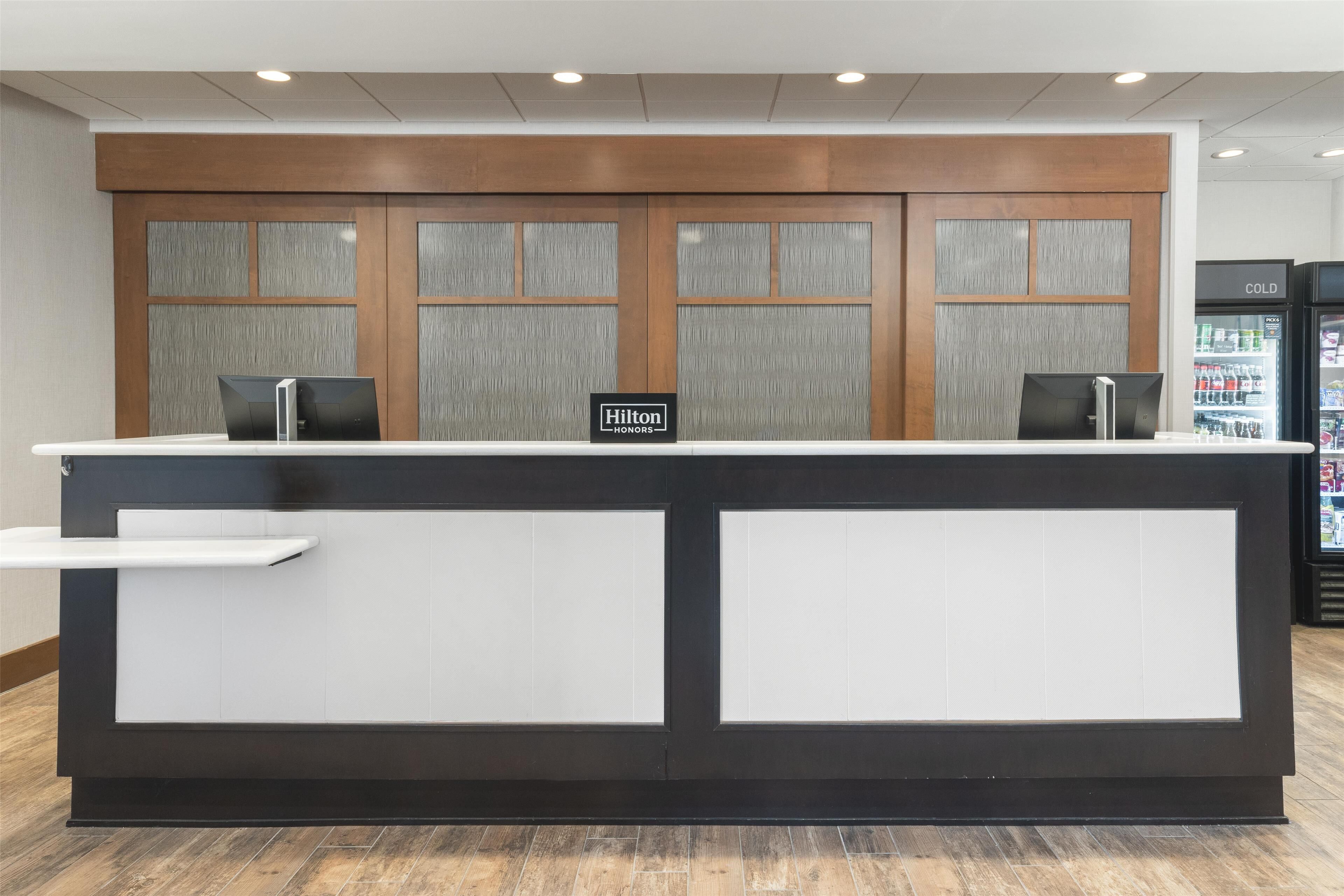 Una mesa de recepción con un cartel que indica Hilton Honors, un mostrador con una decoración blanco y negro, y un refrigerador lleno de bebidas al fondo.