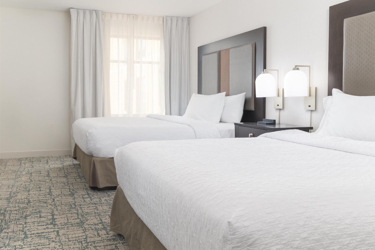 Habitación de hotel con dos camas, un ventanal con cortinas, una mesita de noche con lámpara y un espejo instalado en la pared.