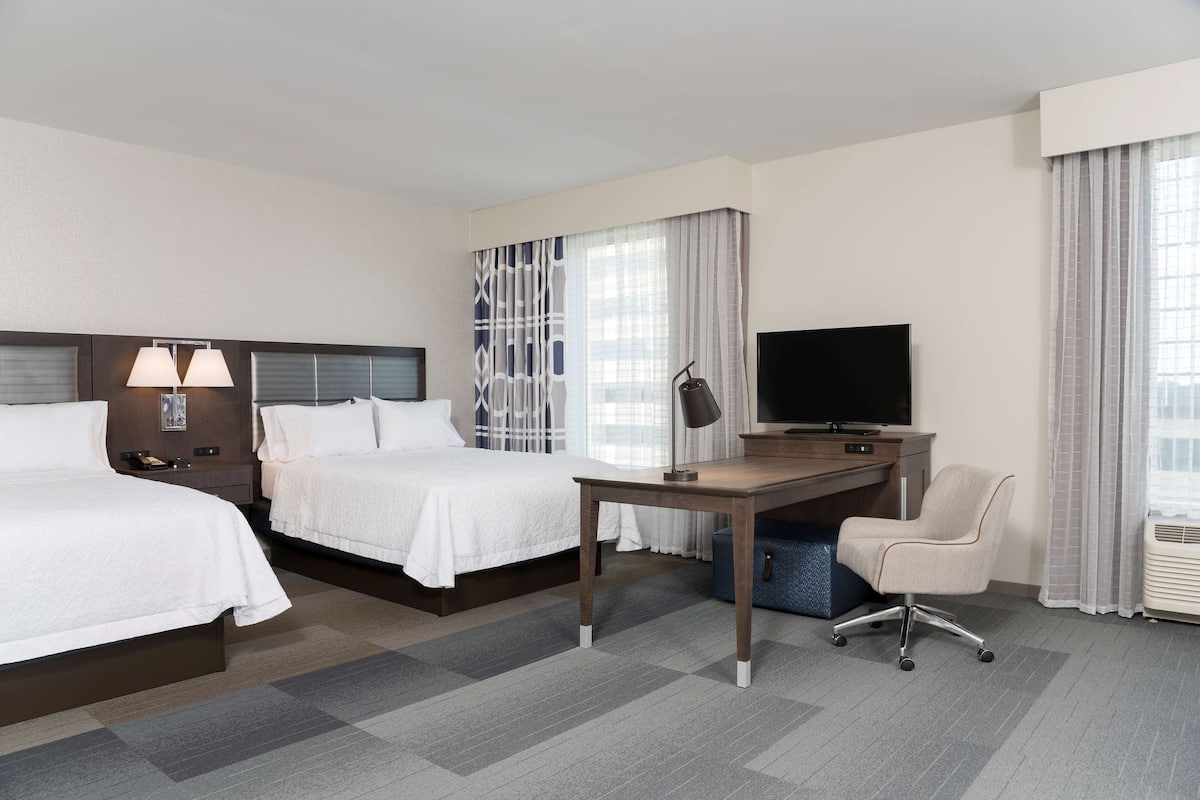 A hotel room with two beds, a desk, a chair, a TV, and a window with curtains.