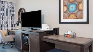 A hotel room with a desk, a flat-screen TV, a coffee maker, a framed artwork, and a chair.