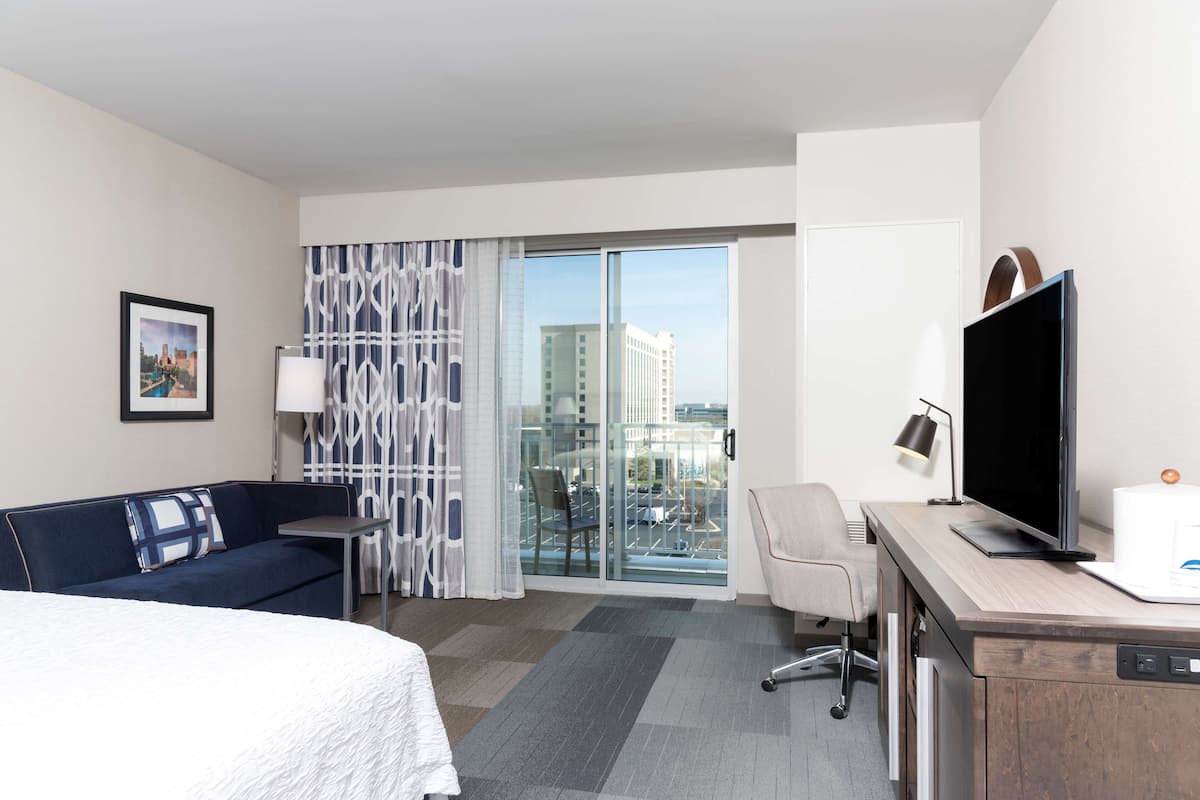 A hotel room with a bed, a desk with a TV, a chair, a sofa, a window with a view of buildings, and a curtain.