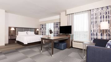 A hotel room with a bed, desk, chair, TV, and large windows with curtains.