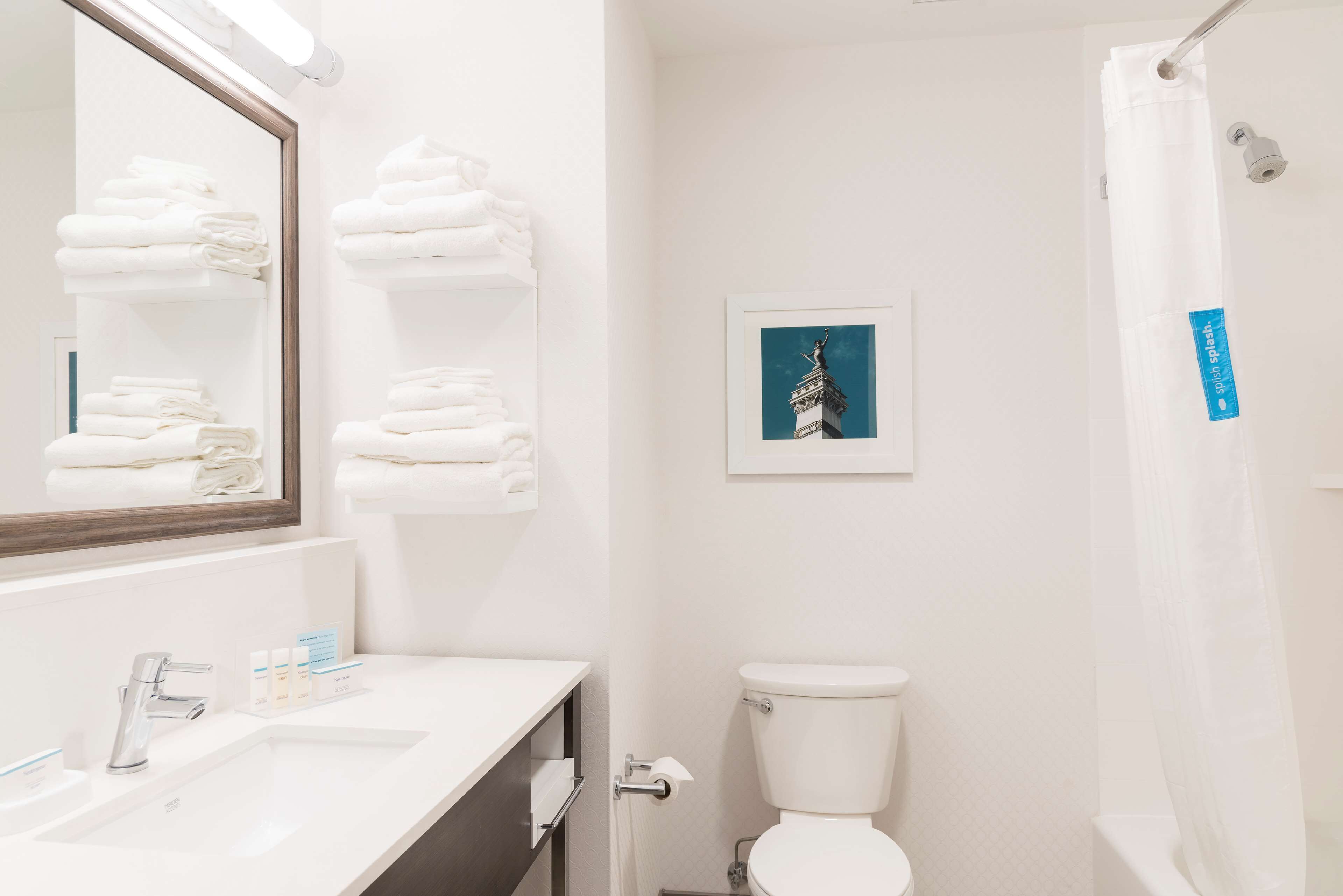 A bathroom with a white sink, mirror, and toilet. There are folded towels on a shelf and a framed picture on the wall.