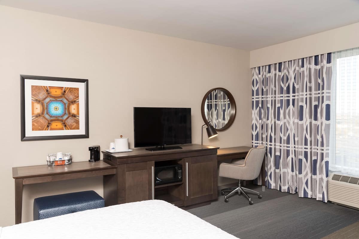 A hotel room with a bed, a desk with a flat-screen TV, a coffee maker, a microwave, a chair, and a large window with patterned curtains.