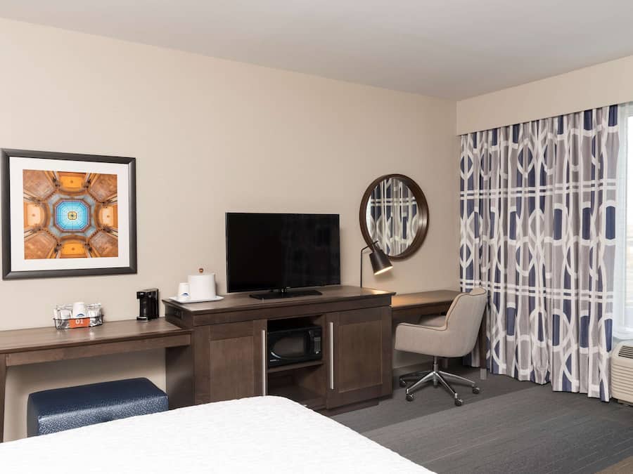 A hotel room with a bed, a desk with a flat-screen TV, a coffee maker, a microwave, a chair, and a large window with patterned curtains.