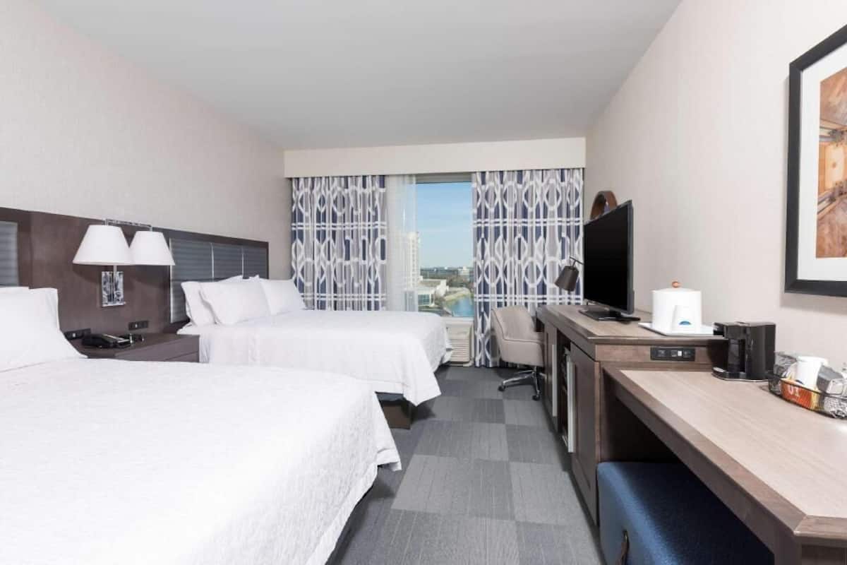Une chambre d’hôtel avec deux lits, un bureau, une chaise, une télévision et une fenêtre avec des rideaux à motifs.