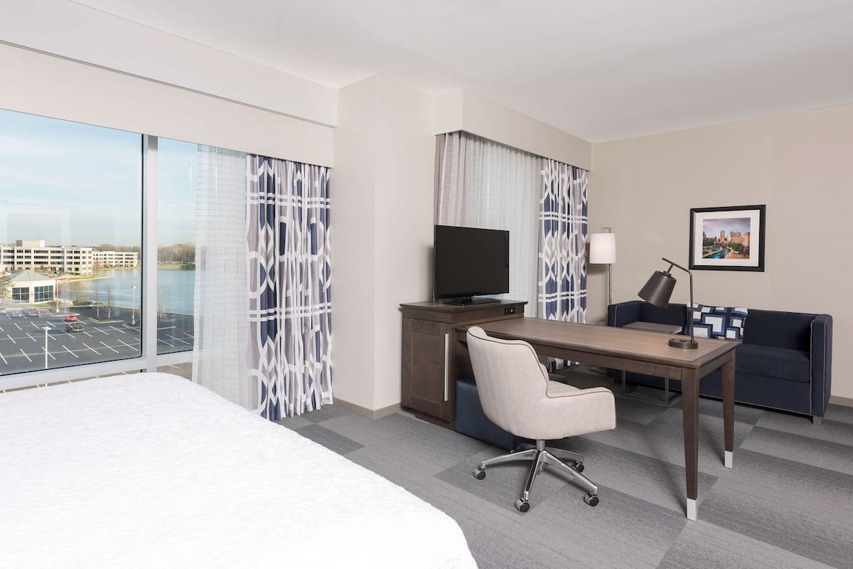 A hotel room with a large bed, a desk with a chair, a television, a sofa, and a large window with a city view.