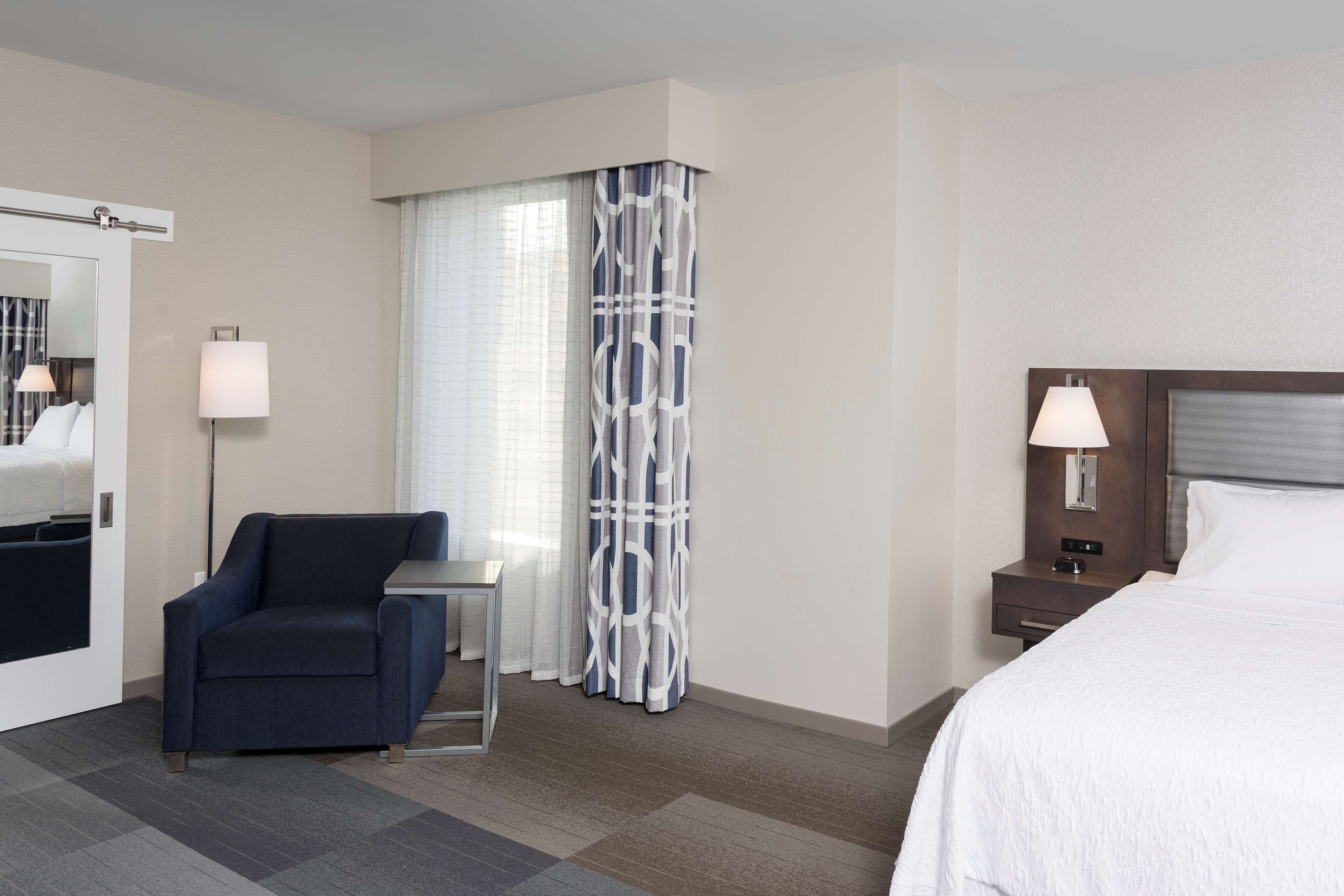 A hotel room with a bed, a chair, a nightstand, a lamp, a mirror, and a window with curtains.