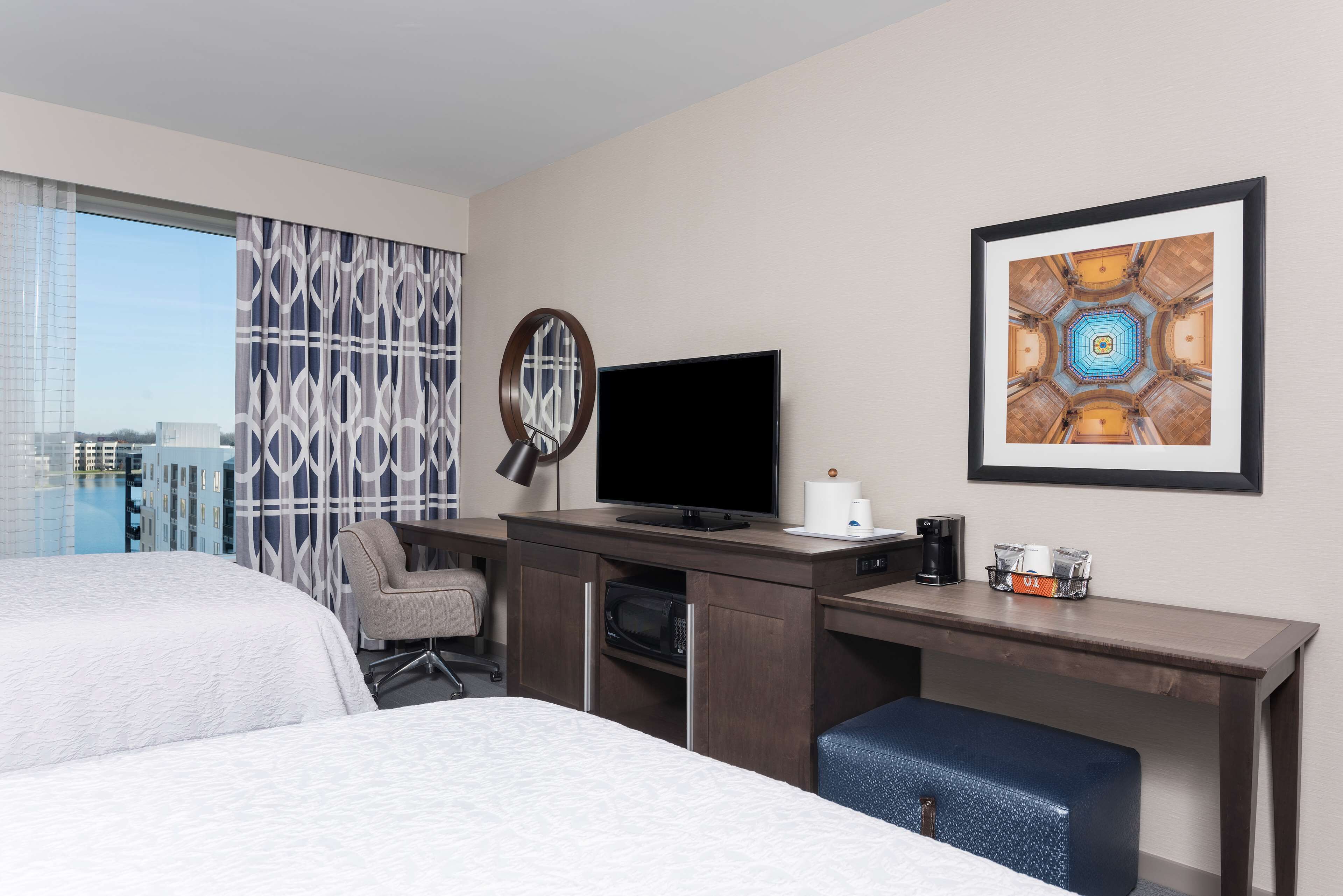 A hotel room with two beds, a desk with a flat-screen TV, a coffee machine, and a framed picture on the wall.