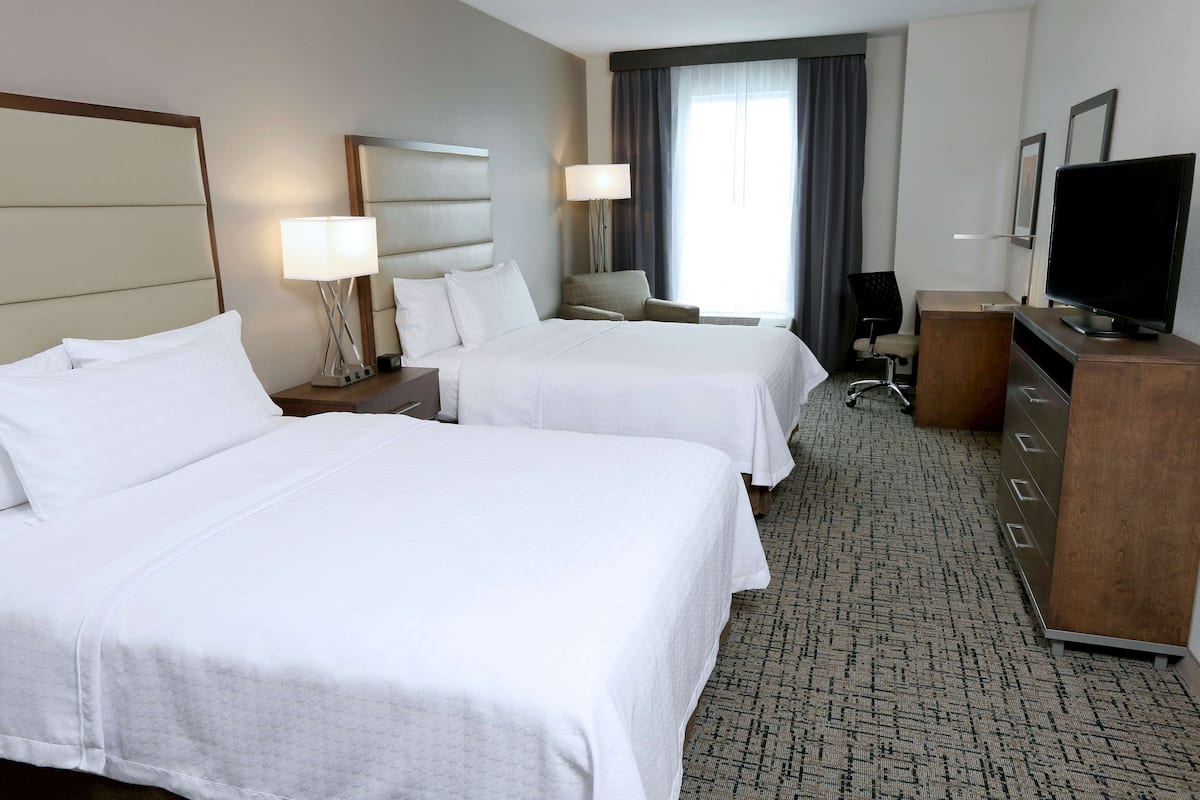 A hotel room with two beds, a desk, a chair, a TV, and a lamp.