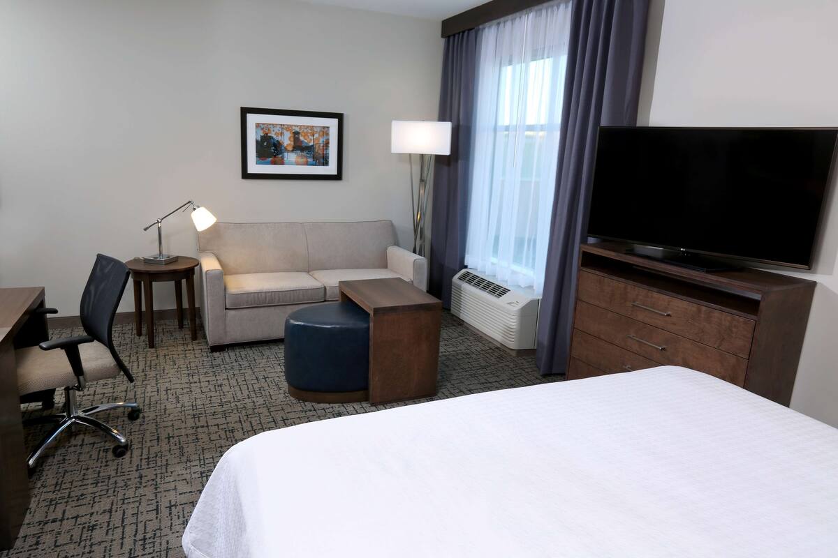 A hotel room with a bed, a desk, a chair, a sofa, a TV, a lamp, a picture on the wall, and a window with curtains.