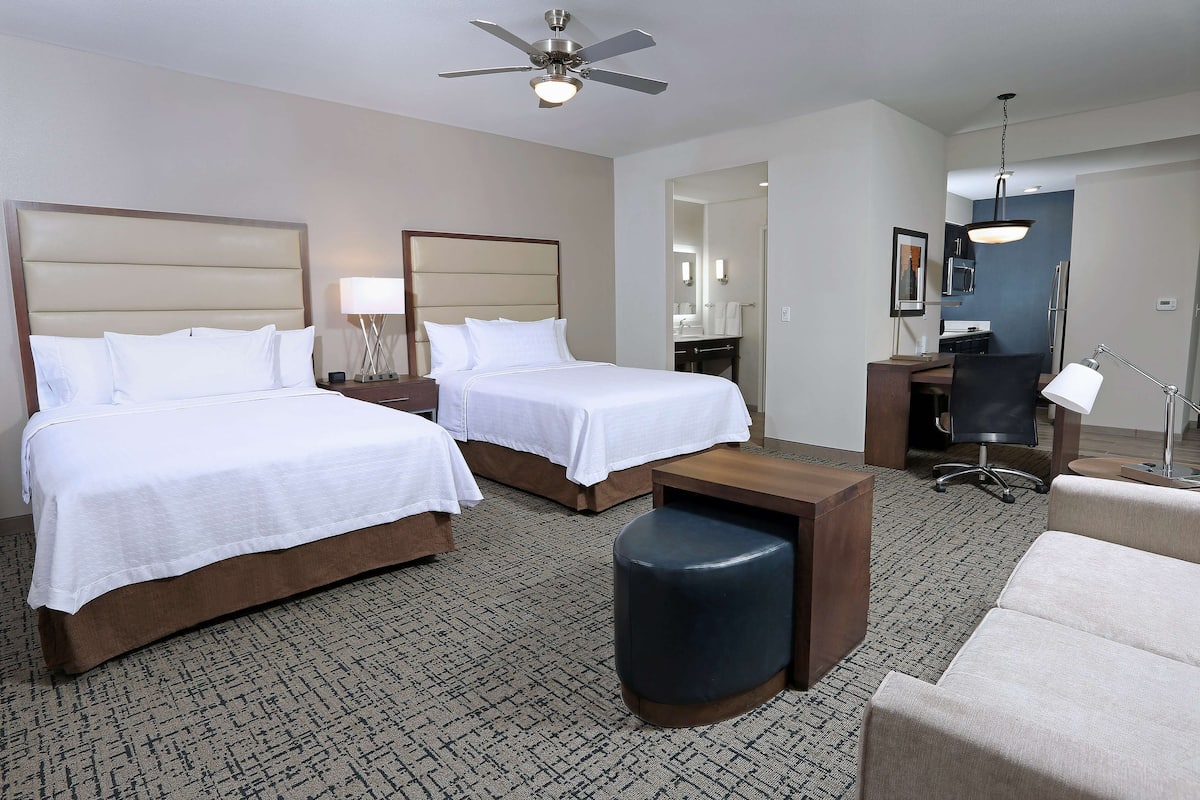 A hotel room with two beds, a desk, a chair, and a ceiling fan.