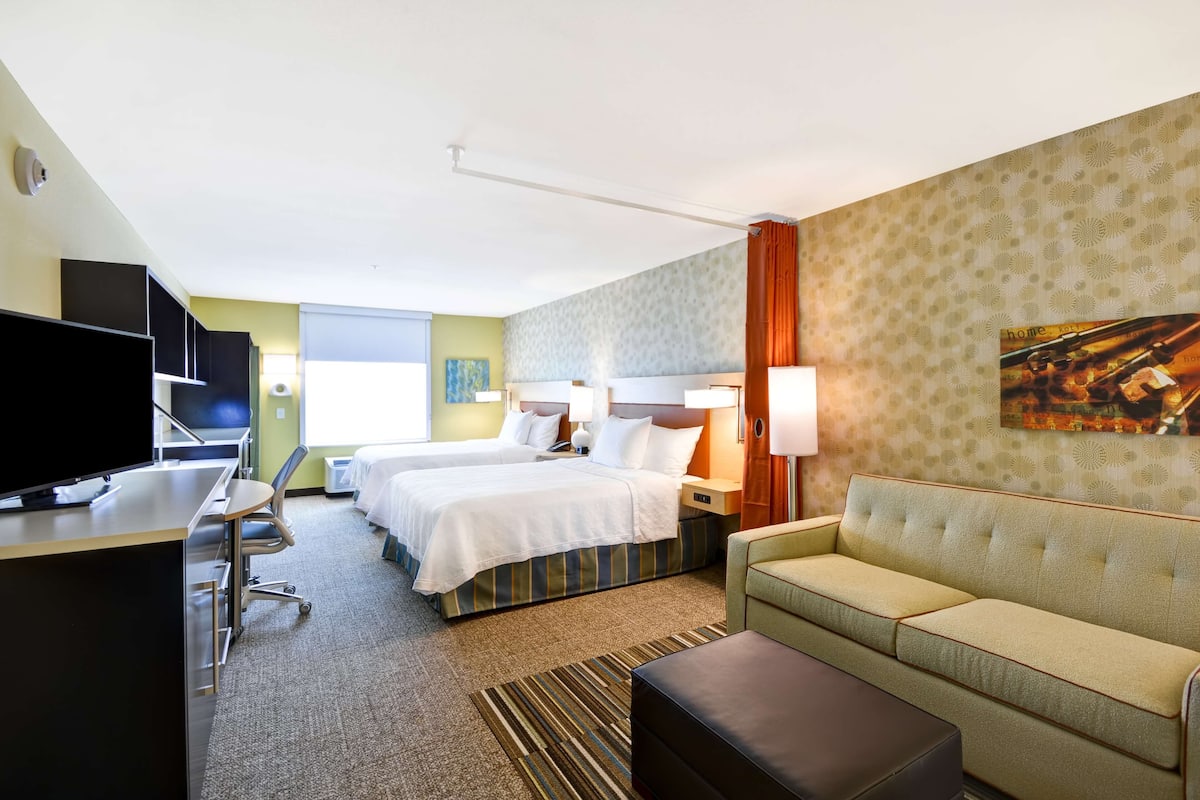 A hotel room with two beds, a sofa, a desk with a TV, and a large window.