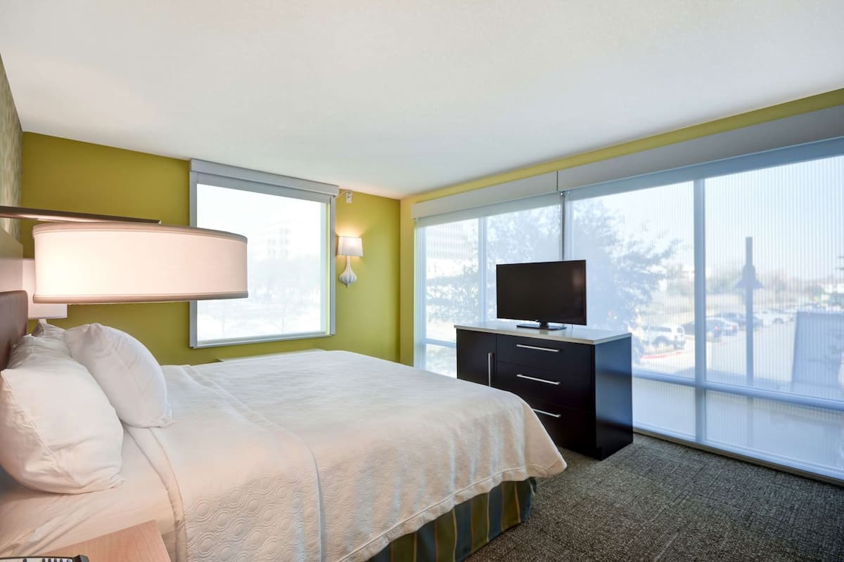 A hotel room with a bed, a TV on a cabinet, and a large window offering a view of the outdoors.