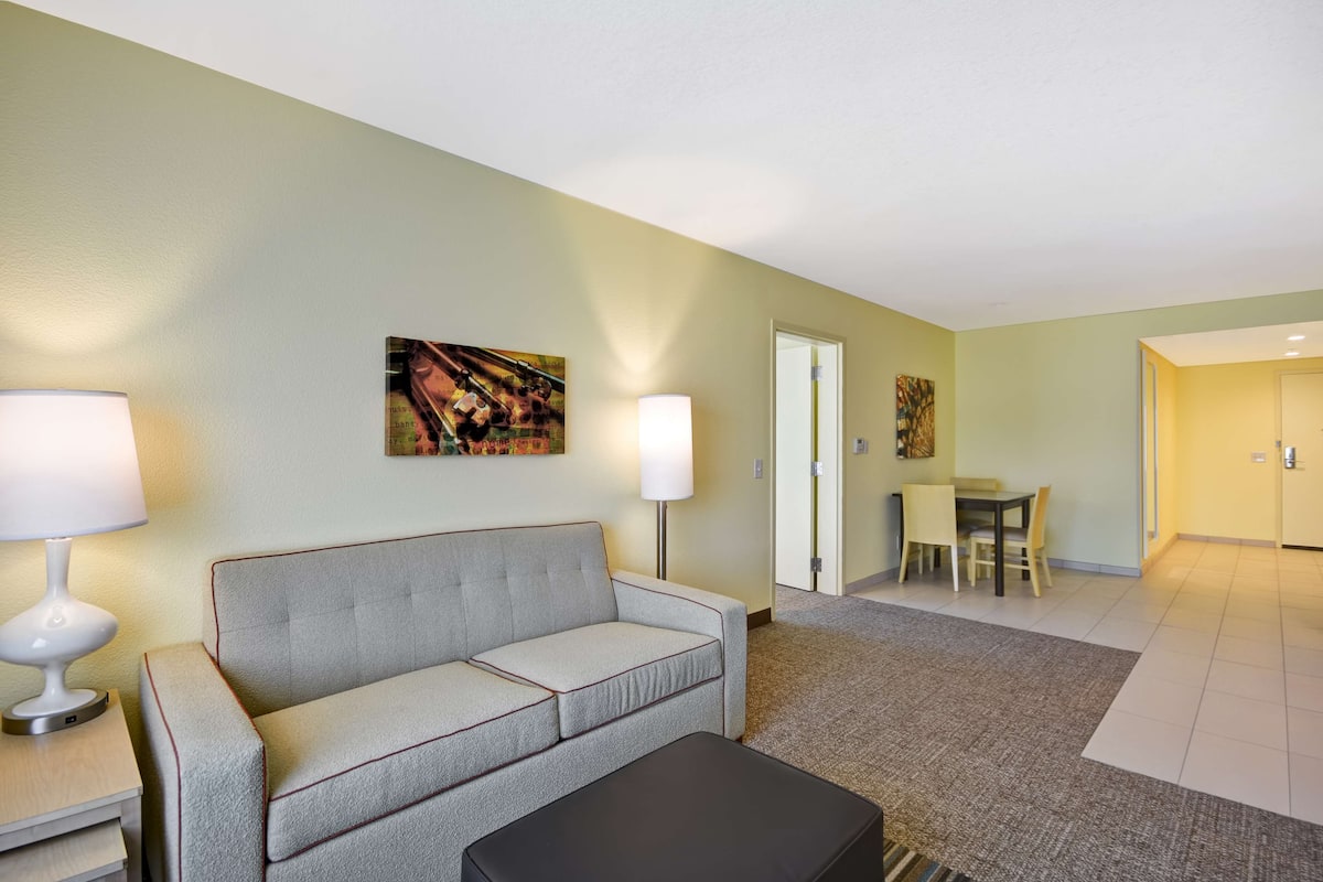 A hotel room with a grey sofa, a black ottoman, a lamp, and a painting on the wall.